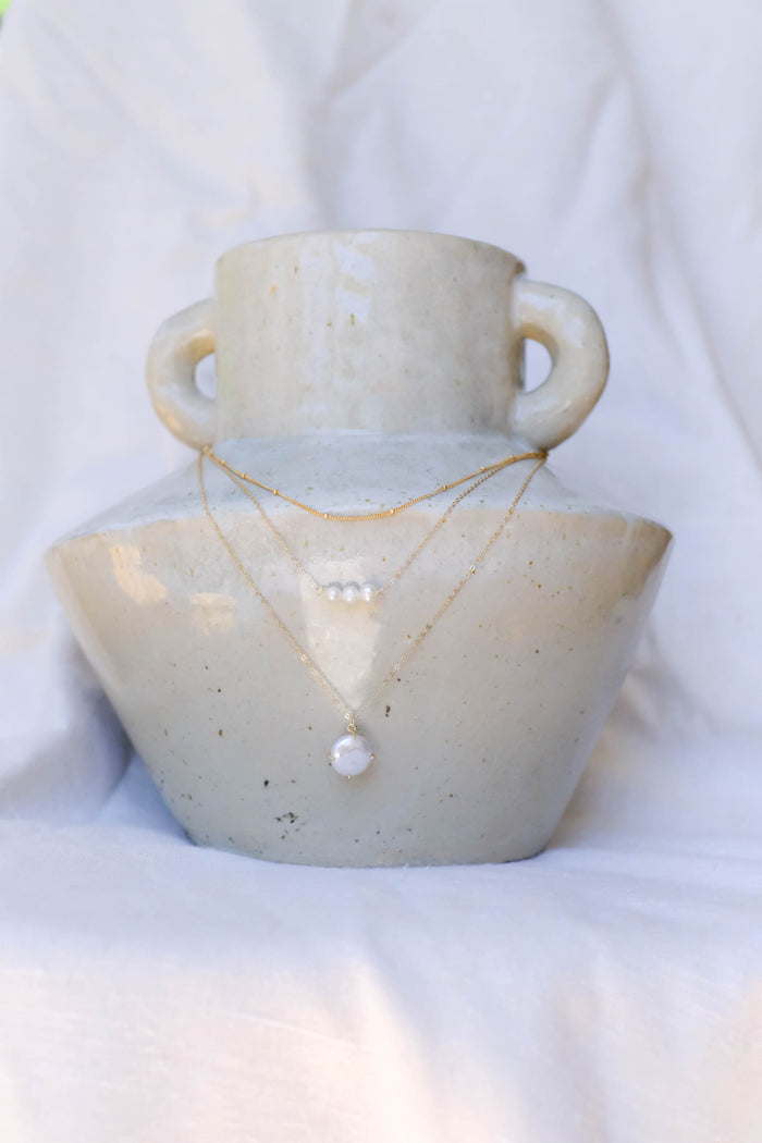 The LLUME Jewelry Pearl Drop Set, featuring a delicate gold chain with a white charm and a pearl pendant necklace, is displayed on a cream ceramic vase against soft white fabric—versatile jewelry to suit any style.