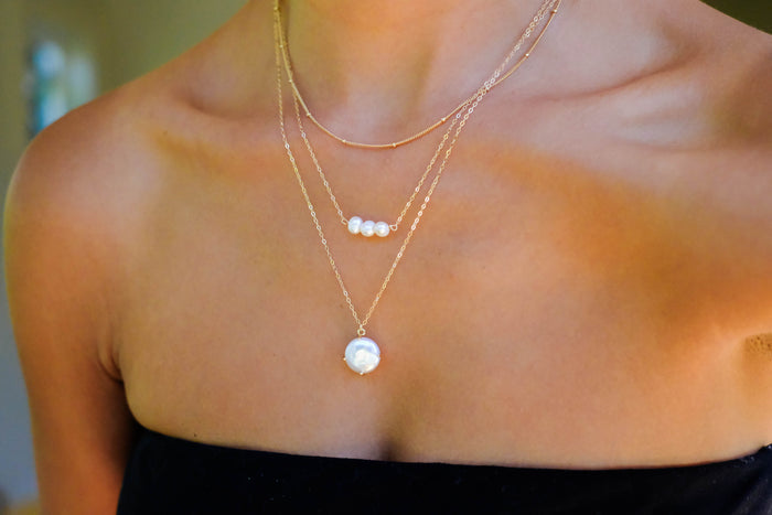 A close-up shows a person wearing the Pearl Drop Set by LLUME Jewelry: one plain chain, one with three small pearls, and a pearl pendant. This versatile set perfectly complements their black strapless top.
