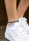 A person wearing a white sneaker has an ankle bracelet with rainbow colors representing the pride flag.