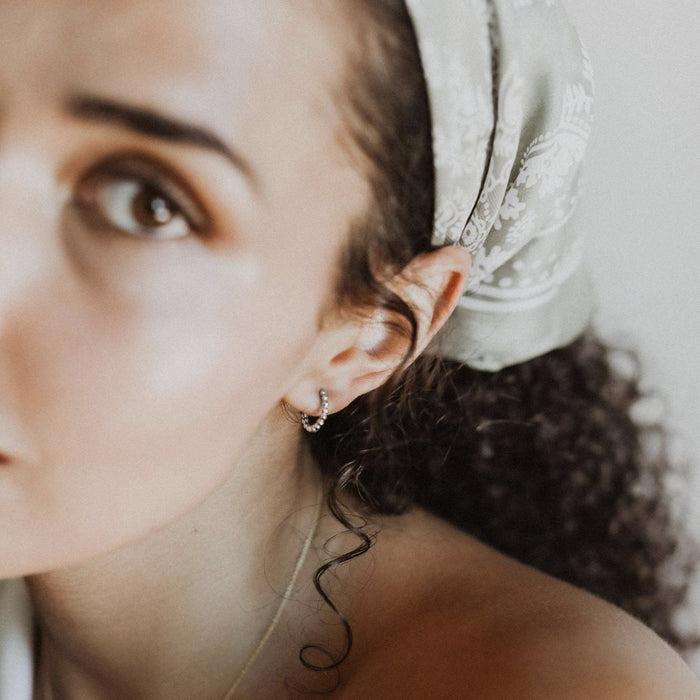 A close-up of a woman with curly hair in a light patterned headscarf wearing LLUME Jewelry Prosecco Beaded Hoops Silver, gazing to the side; only half her face is visible in soft lighting.