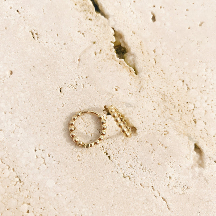 Two gold-plated Prosecco Beaded Hoops by LLUME Jewelry rest on a textured, cream-toned stone surface marked with cracks and small holes.