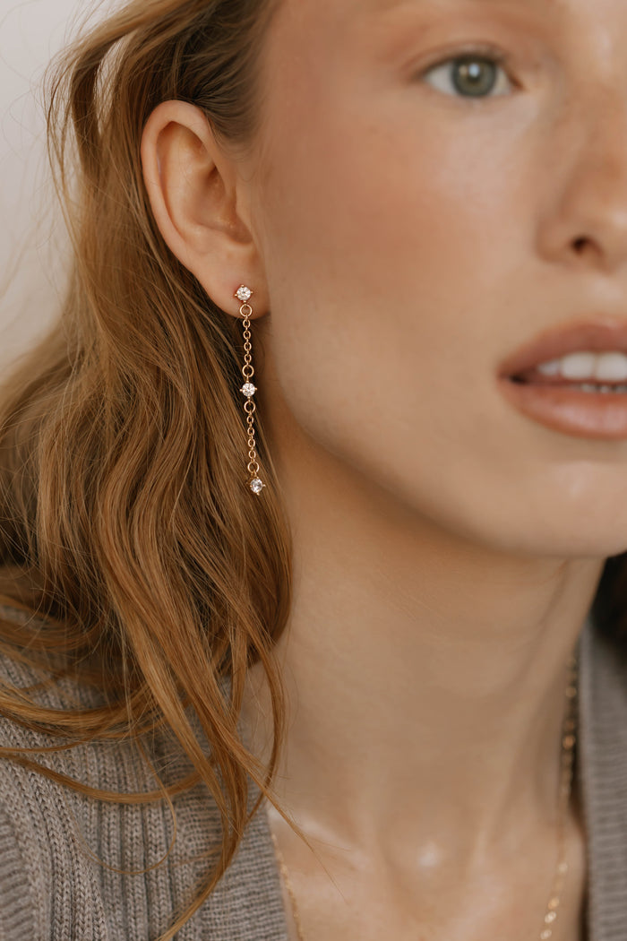 A close-up shows a woman in a grey top wearing LLUME Jewelry's Reverie Earrings—long gold chains with shimmering drops and small gems. Her light brown hair frames her clear skin, with only part of her face and parted lips visible.