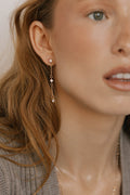 A close-up shows a woman in a grey top wearing LLUME Jewelry's Reverie Earrings—long gold chains with shimmering drops and small gems. Her light brown hair frames her clear skin, with only part of her face and parted lips visible.