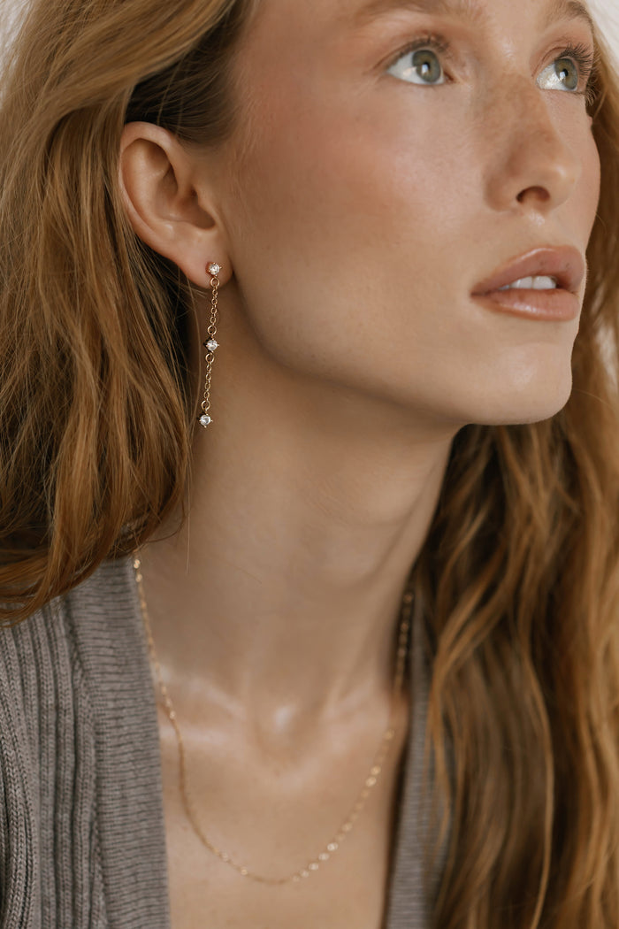 A woman with long light brown hair wears LLUME Jewelry’s shimmering Reverie Earrings and a delicate gold necklace, her subtle makeup and light skin softly illuminated as she gazes upward in a ribbed, taupe-colored top.
