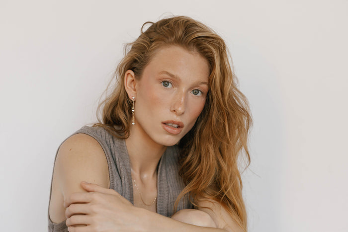 A woman with long, wavy red hair, wearing a sleeveless gray top and LLUME Jewelry’s Reverie Earrings, sits against a white background and gazes neutrally at the camera.