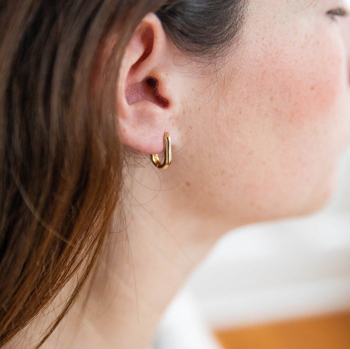 Close-up of a woman's ear with straight brown hair wearing the tarnish-resistant Rigby Earring by LLUME Jewelry. Her light, freckled skin and the softly blurred background highlight the earring's elegant gold-filled design.