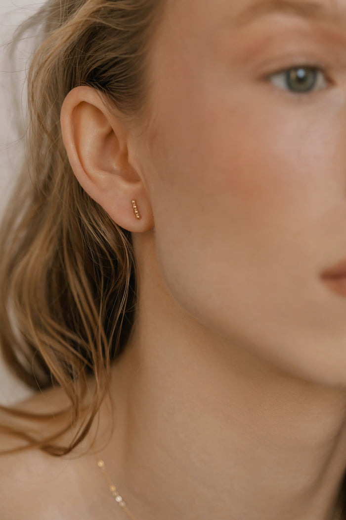 Close-up of a woman wearing LLUME Jewelry's Riley Studs, with wavy blonde hair and smooth skin. The small earring is highlighted against a softly blurred neutral background.