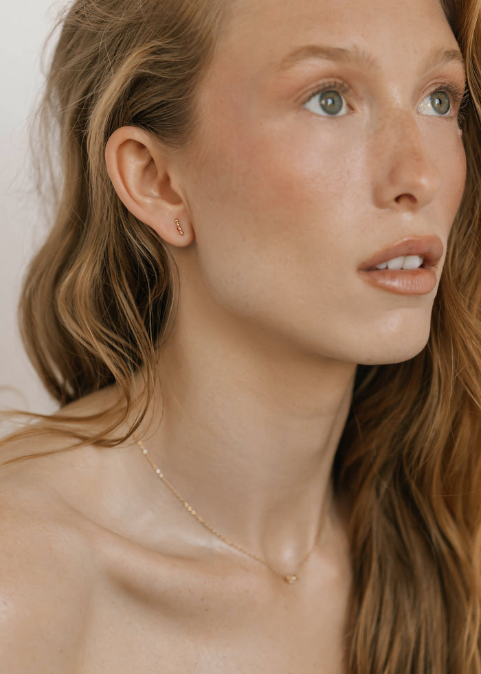 A close-up of a woman with long, wavy light brown hair wearing LLUME Jewelry’s Riley Studs and a delicate gold necklace. Her fair skin glows with natural makeup, highlighting the modern elegance of these everyday earrings.