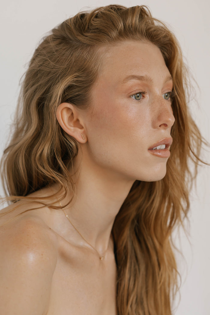 A woman with long wavy light brown hair and fair skin looks to the side, wearing LLUME Jewelry's Riley Studs—delicate gold earrings that add a modern touch to her look. The background is plain and neutral.