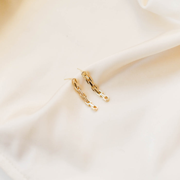 The Sadie Earrings by LLUME Jewelry, featuring 18k gold-plated chain links, rest on a softly draped cream-colored fabric background.