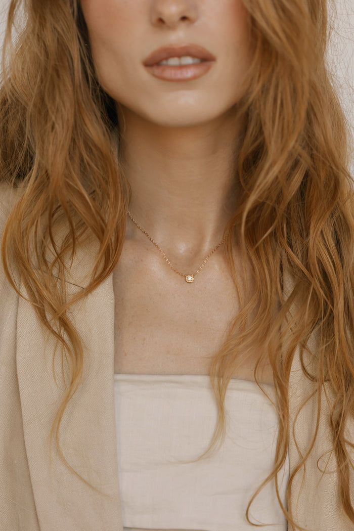 A woman with wavy, light brown hair wears a beige blazer and the LLUME Jewelry Sara Necklace—a delicate gold pendant adding timeless elegance. The cropped image highlights her neck and this classic everyday piece.