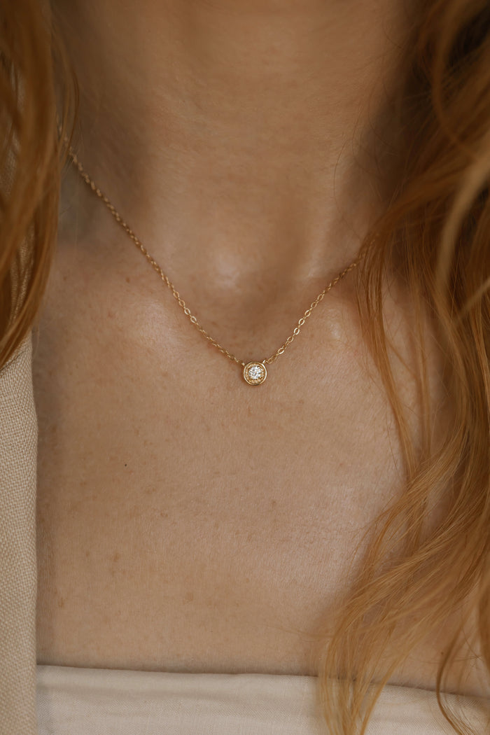 A close-up of a woman wearing LLUME Jewelry's Sara Necklace—a delicate gold chain with a small round pendant and clear gemstone. Wavy auburn hair and neutral clothing frame this timelessly elegant accessory.