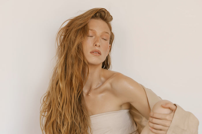 A woman with long, wavy red hair poses against a plain light background, eyes closed and head slightly tilted. She wears a beige off-shoulder top and the Sara Necklace by LLUME Jewelry, exuding timeless elegance in her soft, relaxed look.