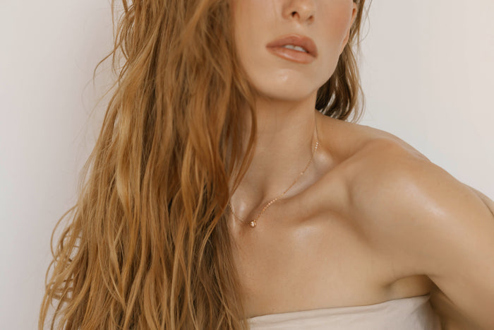 A woman with long, wavy, light brown hair poses in a neutral strapless top against a light background, wearing the Sara Necklace by LLUME Jewelry—a delicate gold piece. Her face is partially visible and shoulders are bare.