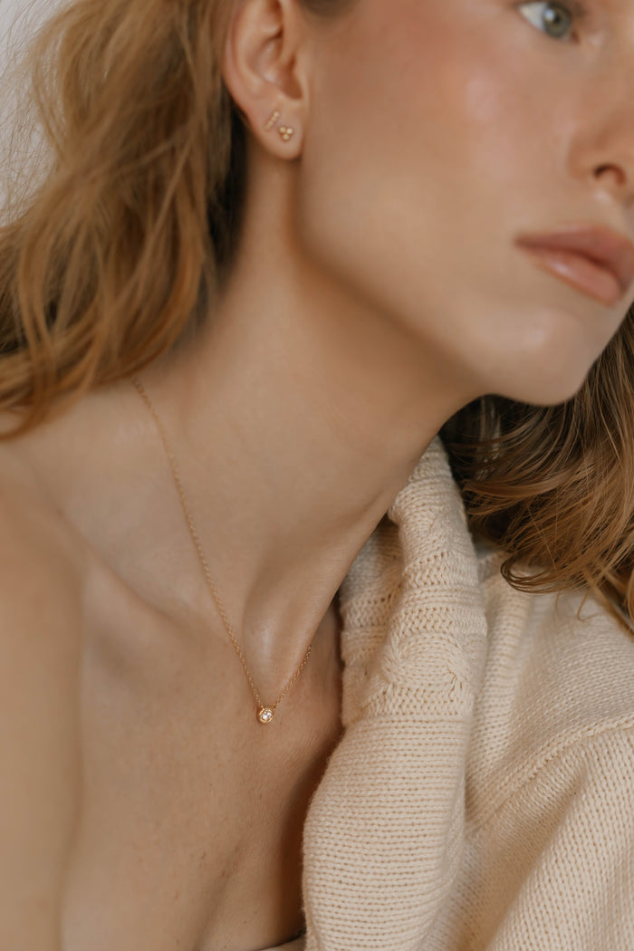 A woman with wavy light brown hair wears the LLUME Jewelry Sara Necklace, a delicate gold piece ideal for daily wear. She pairs it with small gold earrings and a cream knit sweater, creating a look of timeless elegance.