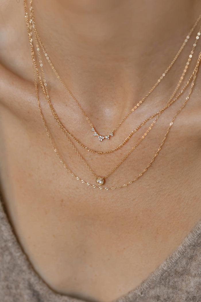 A close-up of a person’s neck wearing three delicate gold necklaces, including the LLUME Jewelry Sara Necklace—each pendant exuding timeless elegance, ideal for daily wear. The person is dressed in a beige top.
