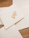 The Sardinia Hoops by LLUME Jewelry—gold earrings with opal drops—rest on cream cards embossed with LLUME, elegantly displayed on a wooden surface.