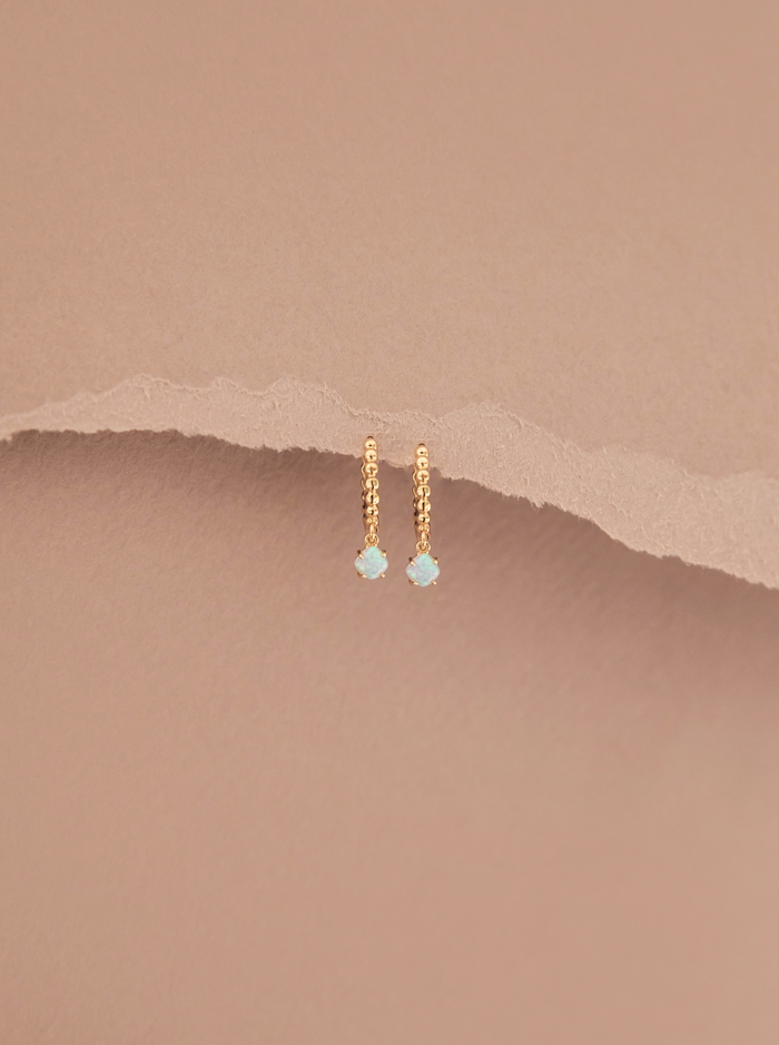 The Sardinia Hoops by LLUME Jewelry—delicate gold hoops with tiny round opal charms—are displayed against a soft beige background with a torn paper texture.