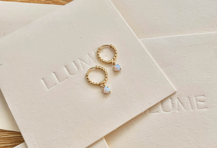 The Sardinia Hoops by LLUME Jewelry feature dainty gold hoops with opal-like stones, elegantly displayed on cream cards embossed with the brand name for a timeless look.