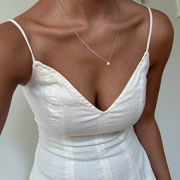 A person wears a sleeveless, V-neck, cream dress with LLUME Jewelry's Sardinia Necklace Silver—a delicate sterling silver piece featuring a white opal charm. The neutral background and the person's head are out of frame.