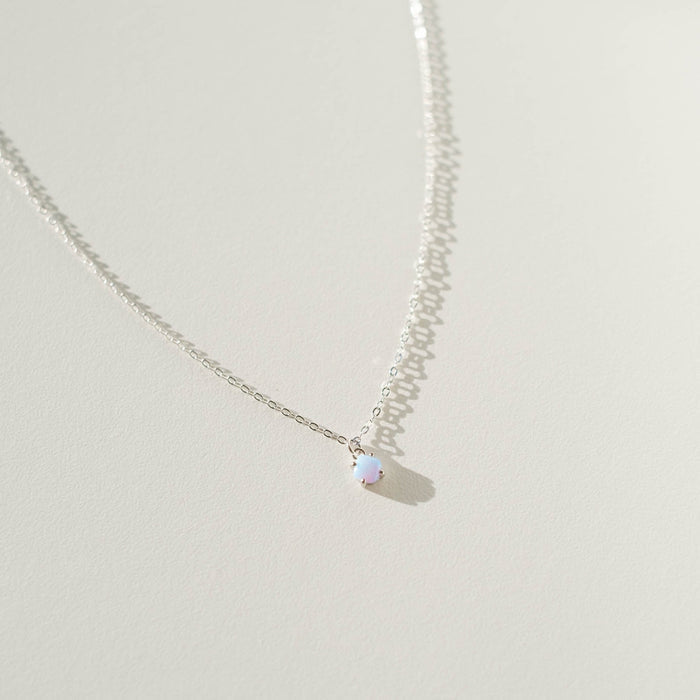 The Sardinia Necklace Silver by LLUME Jewelry, featuring a delicate sterling silver chain and a small round white opal charm, rests on an off-white surface under bright lighting that highlights its elegant simplicity.