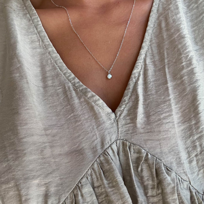 A close-up shows a person wearing the LLUME Jewelry Sardinia Necklace Silver—a sterling silver piece with a small round white opal charm—paired with a light beige, V-neck ruched blouse, highlighting the necklace and fabric texture.