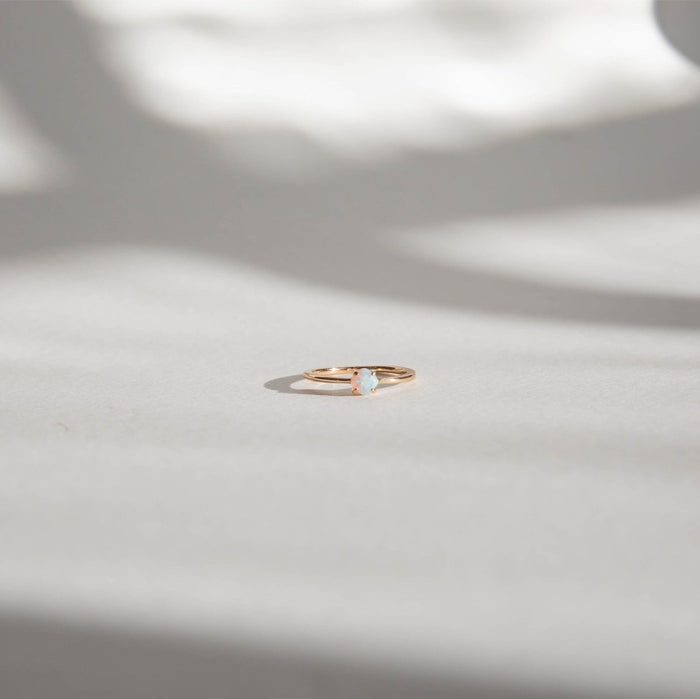 The Sardinia Ring by LLUME Jewelry, a delicate 14k gold filled opal ring, rests on a softly lit white surface with blurred shadows in the background.
