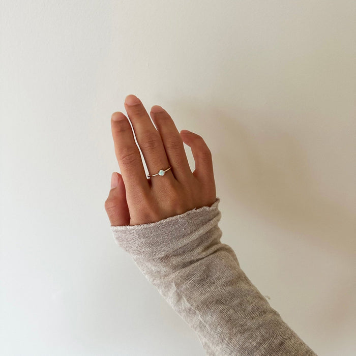 A left hand with a light skin tone, partially covered by a beige long sleeve, displays the Sardinia Ring from LLUME Jewelry against an off-white wall.