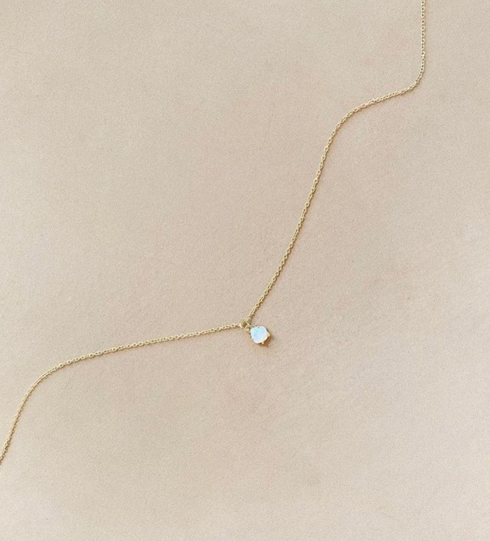 The Sardinia Set by LLUME Jewelry features a delicate gold chain with a small round opal pendant, displayed on a light beige surface—an elegant addition to any jewelry collection.
