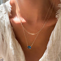 A person wearing a white textured top and the LLUME Jewelry Seacove Daily Set, which features two gold necklaces—one with a thin chain and the other showcasing the striking blue rectangular Seacove Pendant—is shown in natural sunlight.