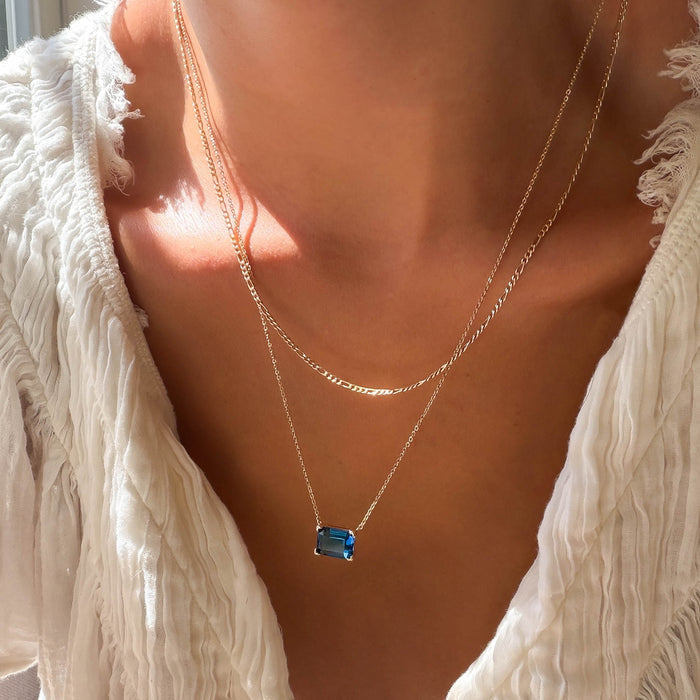 A person wearing a white textured top and the LLUME Jewelry Seacove Daily Set, which features two gold necklaces—one with a thin chain and the other showcasing the striking blue rectangular Seacove Pendant—is shown in natural sunlight.