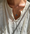 A person wearing a cream-colored, lacy blouse and LLUME Jewelry's Seacove Necklace—a silver multilength chain featuring a rectangular sapphire-blue pendant—shown in natural sunlight.