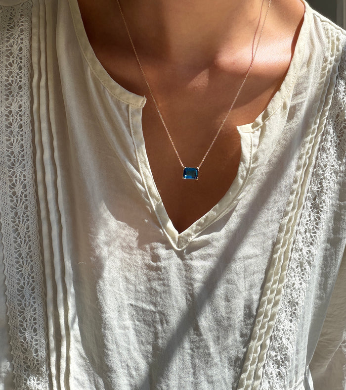 A person wearing a cream-colored, lacy blouse and LLUME Jewelry's Seacove Necklace—a silver multilength chain featuring a rectangular sapphire-blue pendant—shown in natural sunlight.