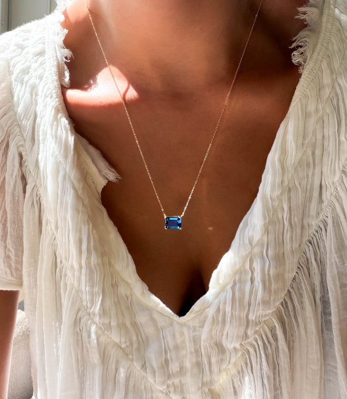 A person wearing a textured white blouse and the LLUME Jewelry Seacove Necklace—a delicate multilength chain featuring a rectangular sapphire-blue pendant—photographed in natural light.