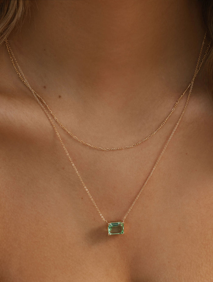A close-up of a woman’s neck wearing the Seafoam Daily Set by LLUME Jewelry: one delicate gold chain is plain, while the other showcases a rectangular green gemstone pendant, capturing effortless coastal elegance.