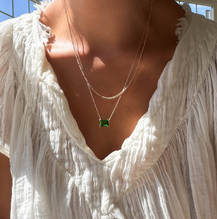 Wearing a crinkled white blouse, a person stands in sunlight near a window, accessorized with the Seagrass Daily Set by LLUME Jewelry—a necklace set featuring two gold necklaces, one adorned with a rectangular green gemstone.