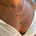 A person wearing the LLUME Jewelry Seagrass Favorites Set—three layered necklaces including a flat chain, a paperclip chain, and a chain with a rectangular green gemstone pendant—all styled over a white, slightly torn top.