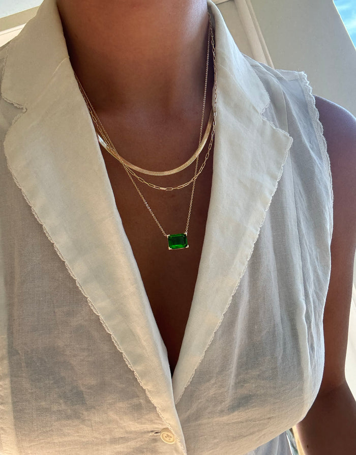Close-up of a person wearing a sleeveless white collared blouse, styled with the LLUME Jewelry Seagrass Favorites Set: three layered gold necklaces, including one with a green rectangular pendant and another featuring an elegant paperclip chain.