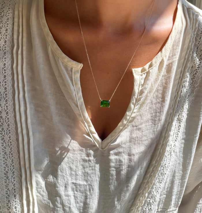 A person wears a white, textured blouse with a v-shaped neckline, accented by LLUME Jewelry’s Seagrass Necklace—a green pendant hanging from a delicate multilength chain. Sunlight casts gentle shadows across the blouse.