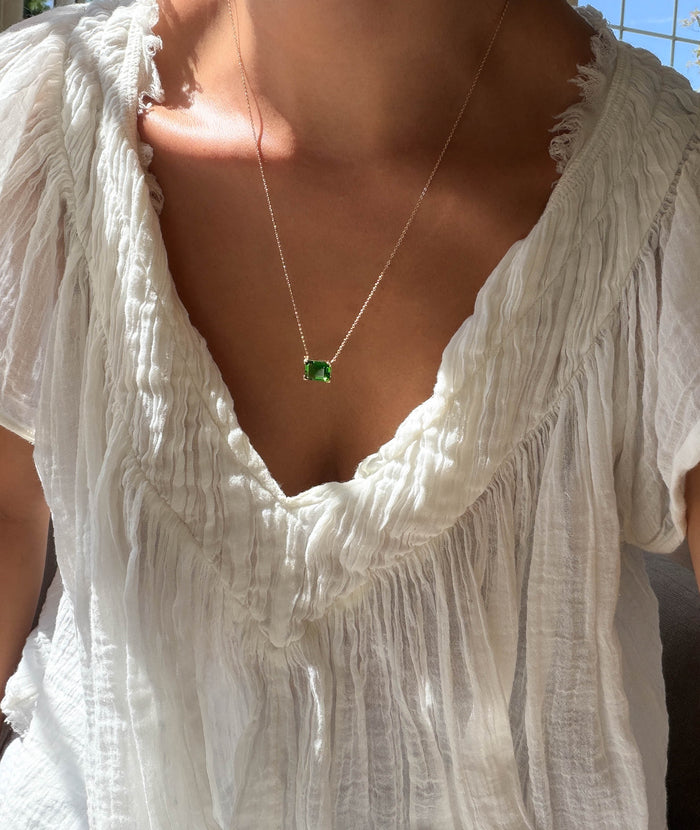 A person wearing a textured white blouse and the LLUME Jewelry Seagrass Necklace—a delicate gold-plated multilength chain with a green rectangular gemstone pendant—shown in natural sunlight.