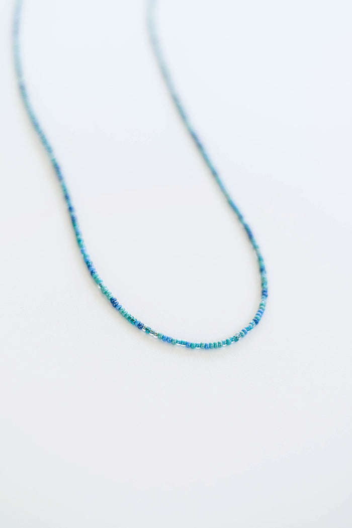 The Searidge Mini Beaded Necklace by LLUME Jewelry features small blue seed beads and is elegantly curved on a white surface, highlighting the charm of handmade jewelry.