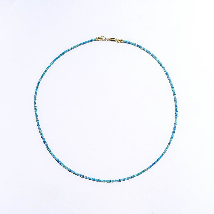 The Searidge Mini Beaded Necklace by LLUME Jewelry features small blue and green seed beads on a thin strand with a gold clasp, displayed in a circle on a plain white background.