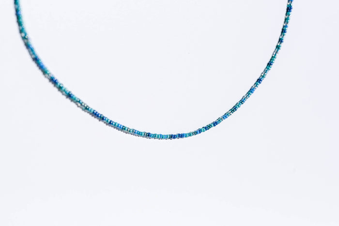 The Searidge Mini Beaded Necklace by LLUME Jewelry features delicate, thin strands of small blue seed beads in varied shades, displayed elegantly against a plain white background.