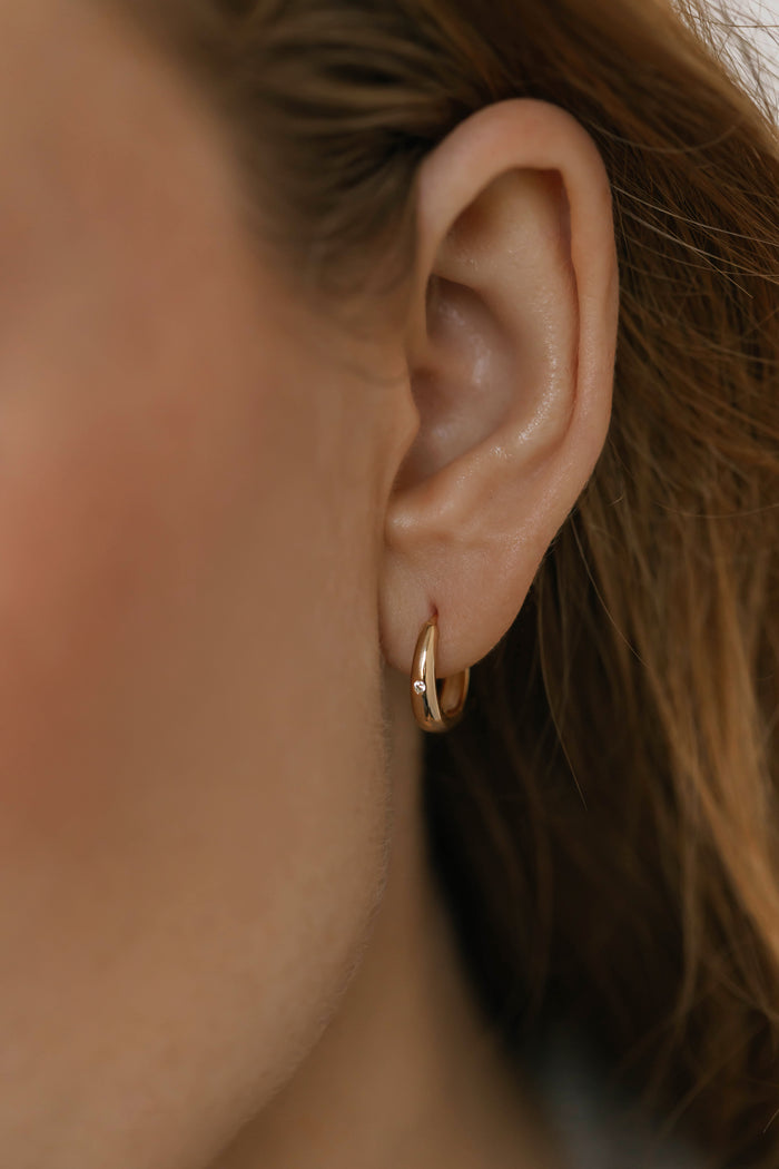 A close-up shows a person with light brown hair wearing LLUME Jewelry's Signature Hoops—classic, lightweight gold earrings that embody timeless elegance—standing out against a softly blurred neutral background.