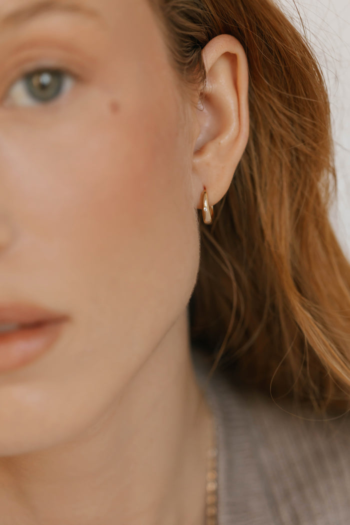 Close-up of a woman with light skin and reddish-brown hair, wearing LLUME Jewelry's Signature Hoops—lightweight earrings known for their timeless elegance. She is dressed in a soft brown top, shown in warm, soft focus.