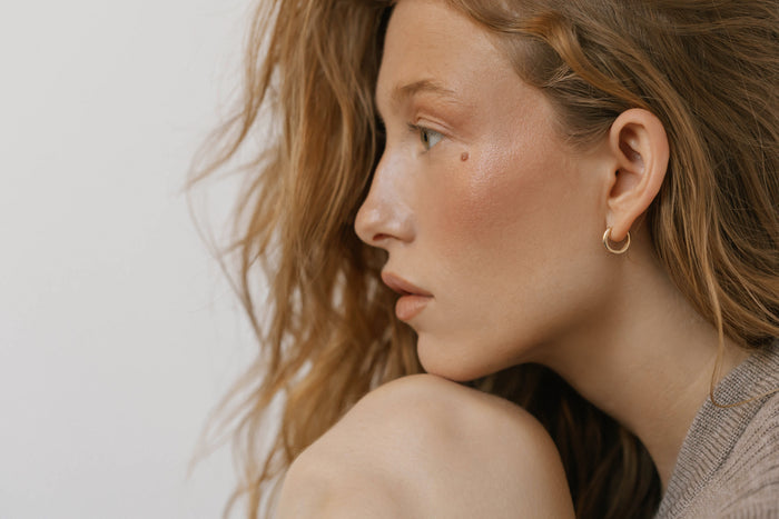 A woman with long, wavy light brown hair and natural makeup sits in profile, gazing thoughtfully to the side. She wears LLUME Jewelry's Signature Hoops, timelessly elegant, paired with a neutral top as her chin rests on her knee.