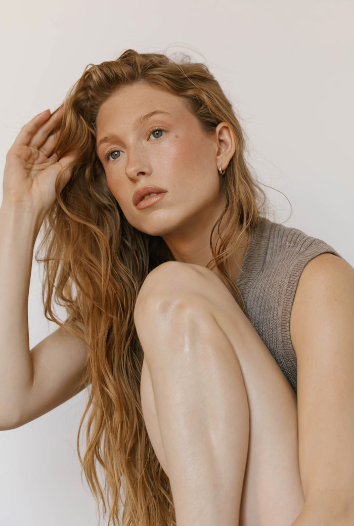 A woman with long wavy blonde hair, in a sleeveless gray top and LLUME Jewelry's Signature Hoops, sits with her knees up against a plain light background, gazing thoughtfully to the side—capturing effortless elegance.