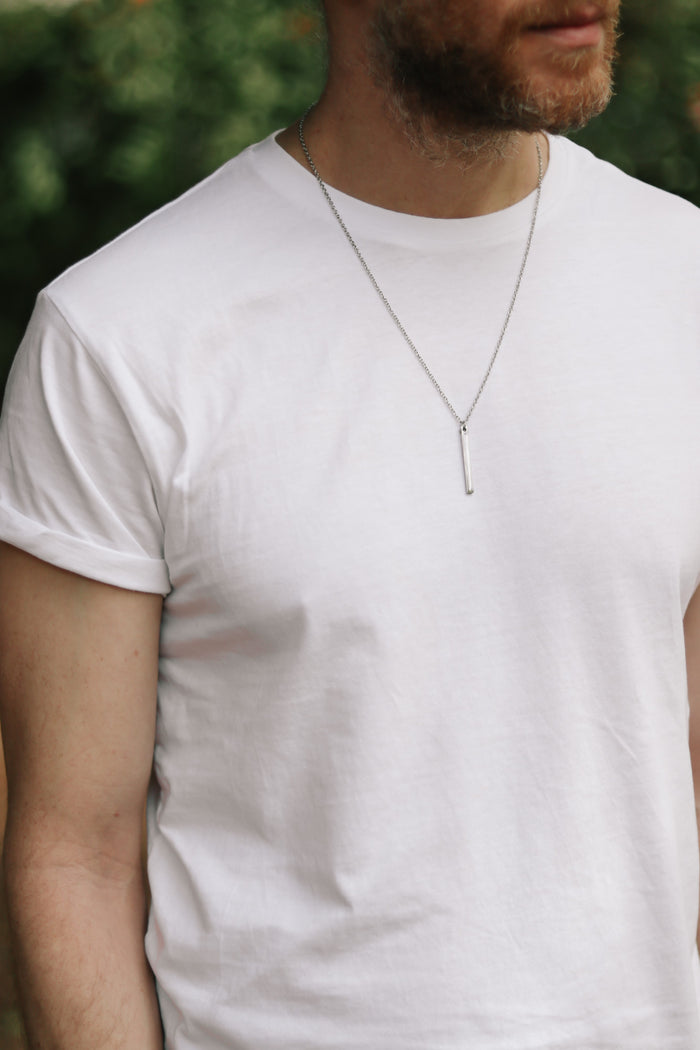 A man wearing a white t-shirt with a silver bar pendant necklace on, which is accompanied by a stainless steel chain.