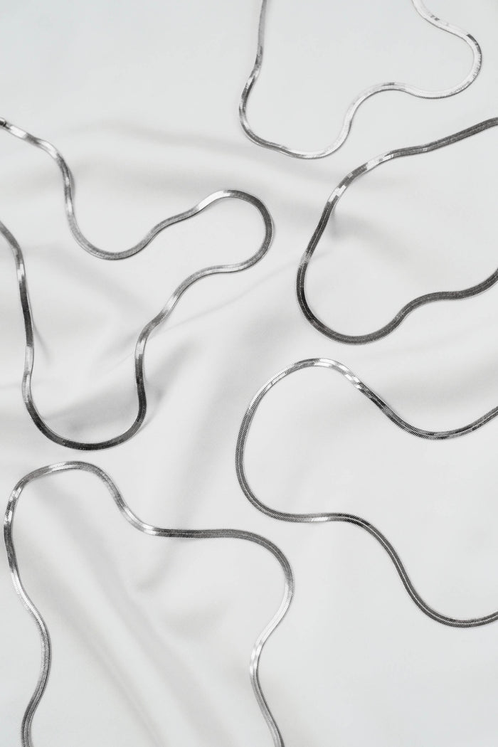 Abstract wavy silver lines on smooth white fabric evoke the elegance of the Skinny Hailey Chain Silver by LLUME Jewelry, with solid 925 silver tones forming organic patterns, soft shadows, and gentle folds in the material.