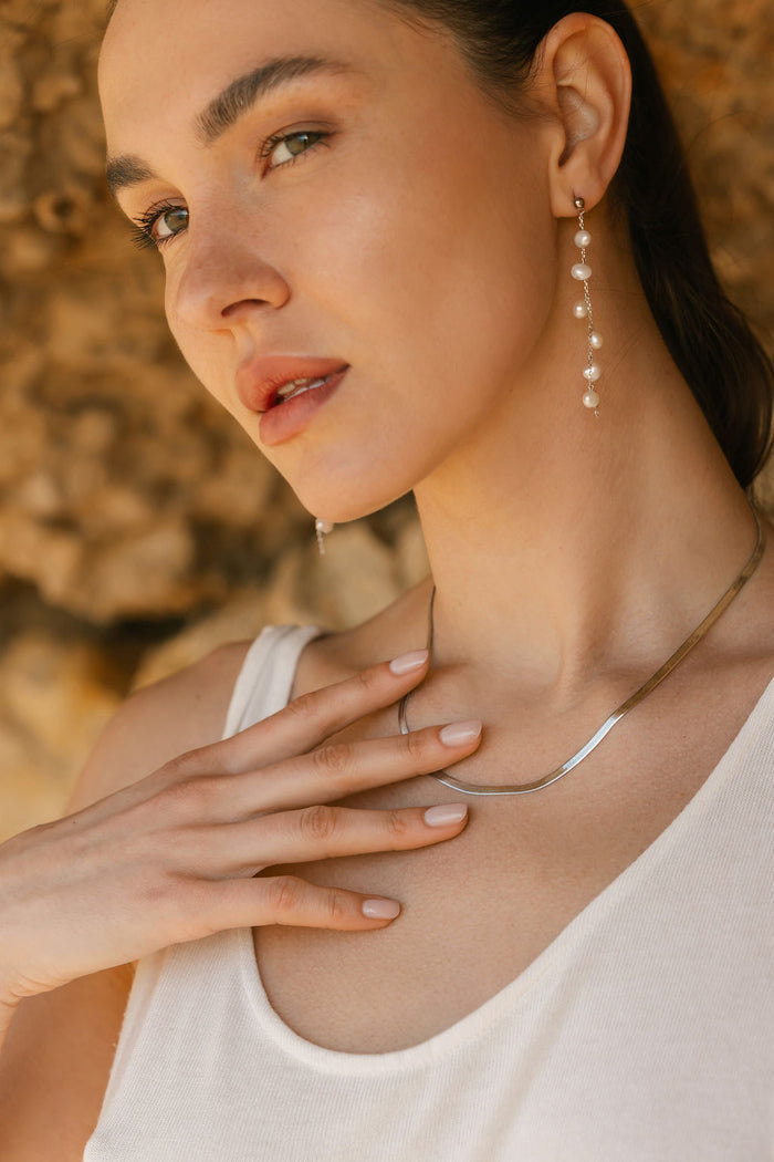 A woman in a sleeveless white top wears long pearl drop earrings and the Skinny Hailey Chain Silver necklace from LLUME Jewelry, gently touching her collarbone against a softly blurred, earthy-toned background.
